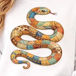 May include: White sweatshirt with a colourful patchwork snake design. The snake is made up of various patterned fabrics in shades of blue, orange, yellow, and brown. The snake's head has an eye and a red tongue.