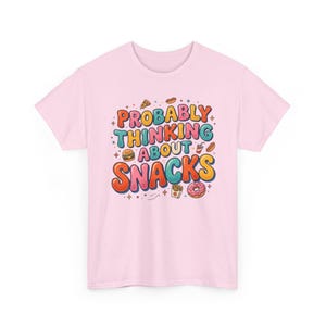 Pode incluir: Camiseta rosa claro com a frase "Probably Thinking About Snacks" em letras coloridas e retrô. O design inclui ilustrações de pizza, hambúrguer, batatas fritas, donut e refrigerante.