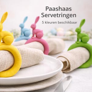Op de afbeelding: Paas-tafelsetting met kleurrijke konijnen servetringen. De ringen zijn geel, blauw, roze en groen, elk rond een opgerolde beige linnen servet. Witte borden en een bloemstuk maken het feestelijke geheel compleet.
