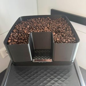 May include: A black plastic coffee bean hopper filled with roasted brown coffee beans. The hopper features a central U-shaped cutout with a small metallic perforated grid at the base, positioned above a textured black surface of a coffee machine.
