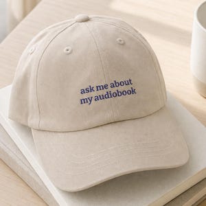 'Ask Me About My Audiobook' dad hat resting on stacked books on a pale desk, styled with wireless earbuds, a ceramic dish, and a matte mug in bright natural morning light.