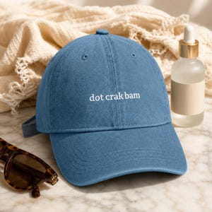 'Dot Crak Bam' embroidered denim mahjong dad hat — adjustable strap, modern aesthetic. 3/4 angle on pale marble with frosted oil bottle and tortoiseshell sunglasses in warm morning light.