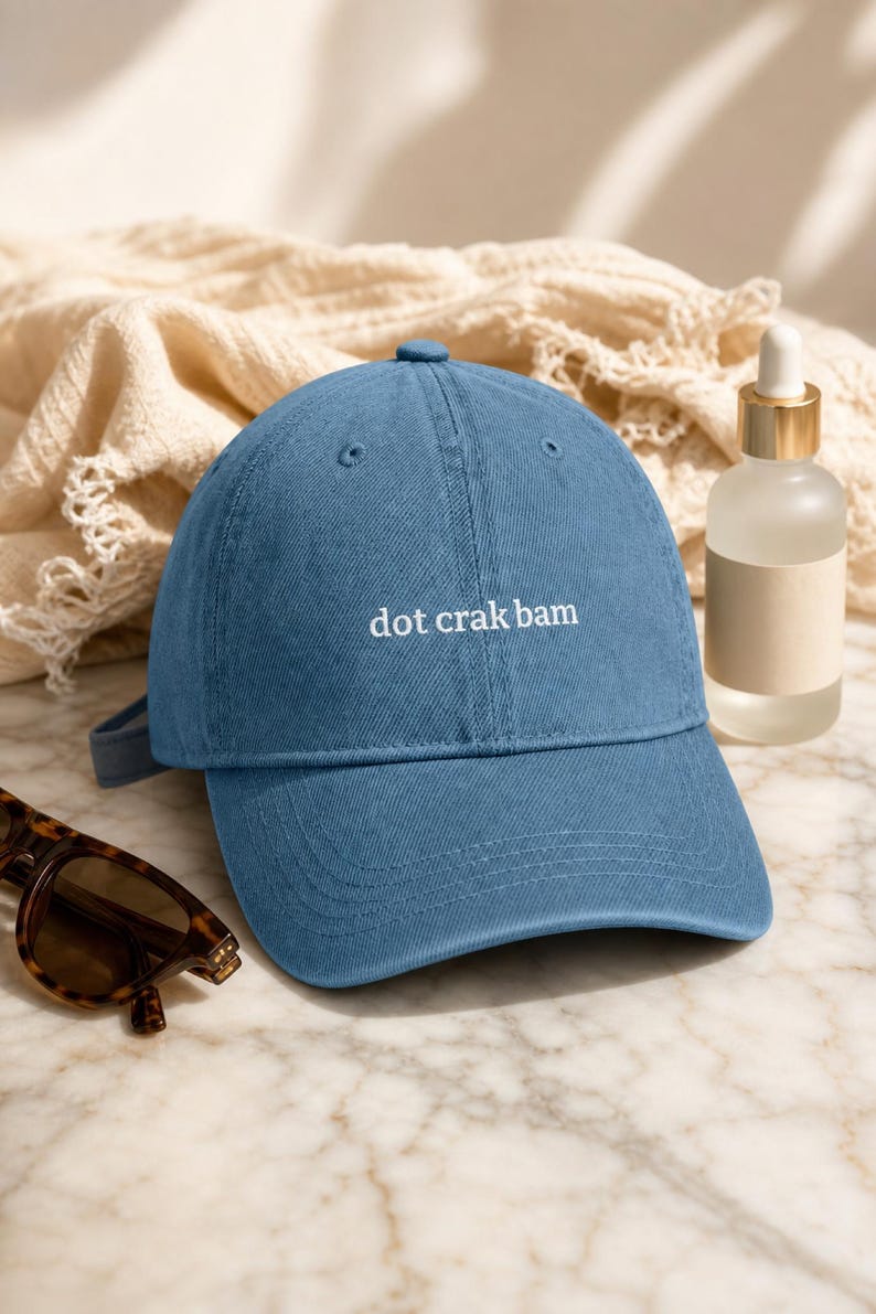 'Dot Crak Bam' embroidered denim mahjong dad hat — adjustable strap, modern aesthetic. 3/4 angle on pale marble with frosted oil bottle and tortoiseshell sunglasses in warm morning light.