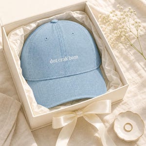 'Dot Crak Bam' embroidered mahjong dad hat in a cream gift box with satin ribbon — denim adjustable cap presented as a mahjong lover gift. Overhead editorial flat lay on cream linen with baby's-breath.