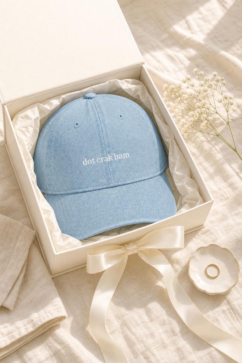 'Dot Crak Bam' embroidered mahjong dad hat in a cream gift box with satin ribbon — denim adjustable cap presented as a mahjong lover gift. Overhead editorial flat lay on cream linen with baby's-breath.