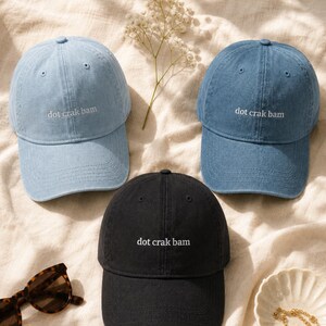 'Dot Crak Bam' embroidered mahjong dad hat in black and light blue — both colorways of the denim adjustable cap shown together. Mahjong lover gift options. Overhead flat lay on cream linen in morning light.