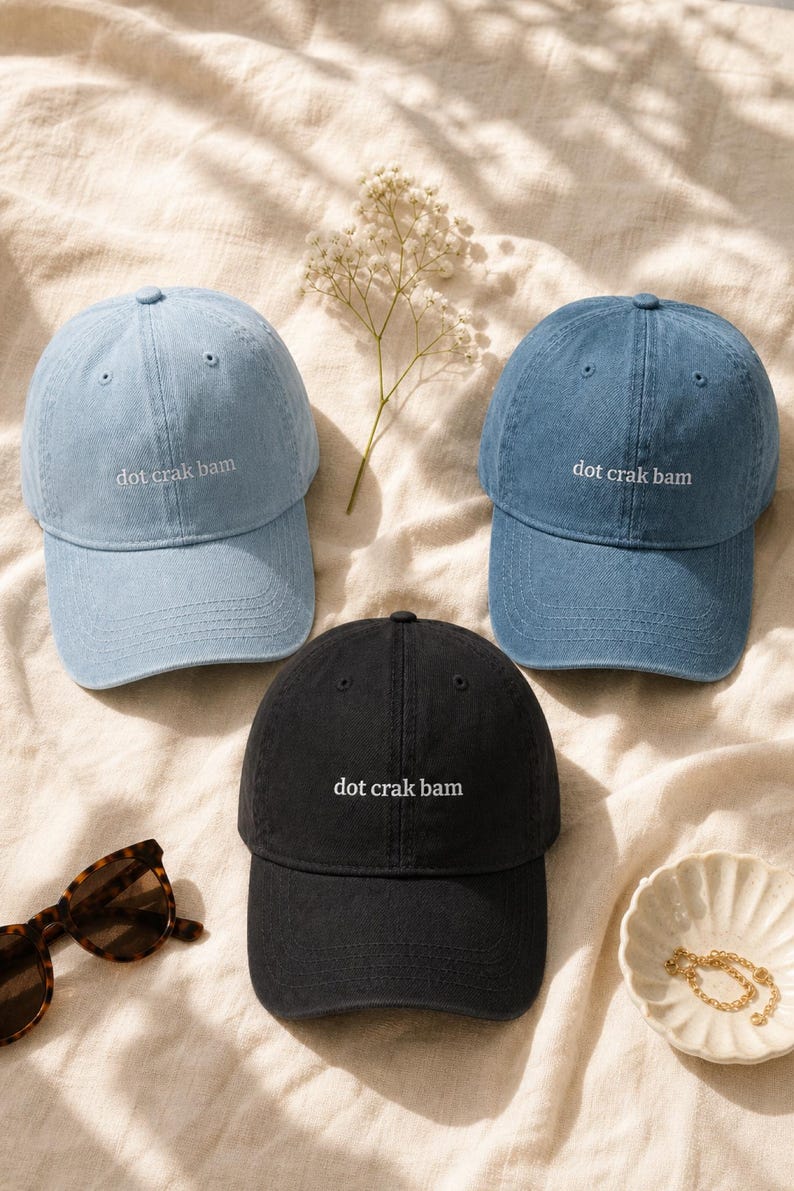 'Dot Crak Bam' embroidered mahjong dad hat in black and light blue — both colorways of the denim adjustable cap shown together. Mahjong lover gift options. Overhead flat lay on cream linen in morning light.
