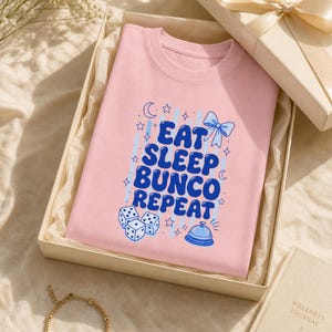 'Eat Sleep Bunco Repeat' bunco dice graphic tee folded in a cream gift box with satin ribbon — bunco lover gift tee styled in an elevated wellness editorial gift flat lay.