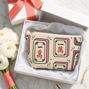 Mahjong tile-print accessory pouch in a glossy white gift box with coral grosgrain ribbon — mahjong lover gift presentation on white marble with ranunculus and cowrie shells.