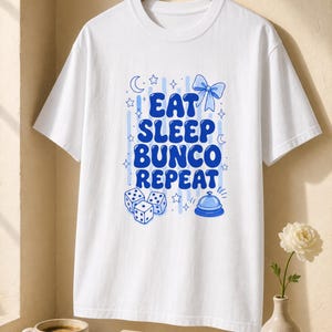 'Eat Sleep Bunco Repeat' bunco dice graphic tee on a wood hanger in a warm editorial wellness setting — bunco group tee lifestyle gift, chest print sharp and legible, styled hanger shot.