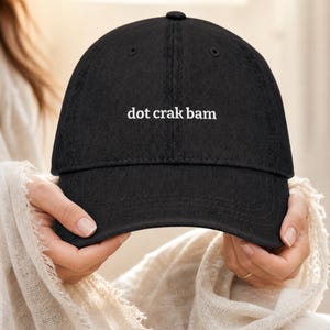 'Dot Crak Bam' embroidered mahjong denim dad hat held gently in hands showing front embroidery — adjustable strap cap, mahjong lover gift. Warm window light editorial close-up with gold ring detail.