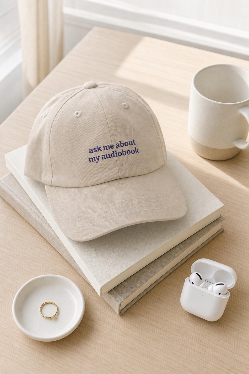 'Ask Me About My Audiobook' dad hat resting on stacked books on a pale desk, styled with wireless earbuds, a ceramic dish, and a matte mug in bright natural morning light.