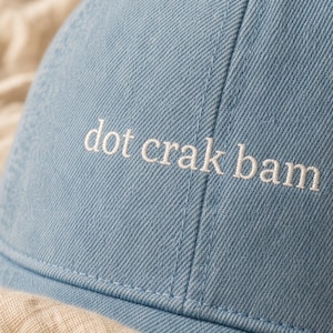 'Dot Crak Bam' embroidered mahjong hat — close-up macro detail of stitching on a denim dad cap. Perfect mahjong lover gift. Warm morning light reveals thread texture on cream linen.