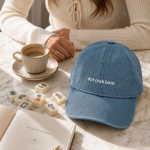 'Dot Crak Bam' embroidered mahjong dad hat styled at a sunlit marble table with mahjong tiles and espresso — denim adjustable cap, ideal mahjong player gift. Warm morning editorial lifestyle scene.