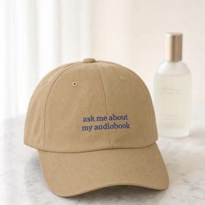 'Ask Me About My Audiobook' embroidered dad hat styled on pale marble beside a frosted glass bottle, shot in bright airy window light with shallow depth of field.