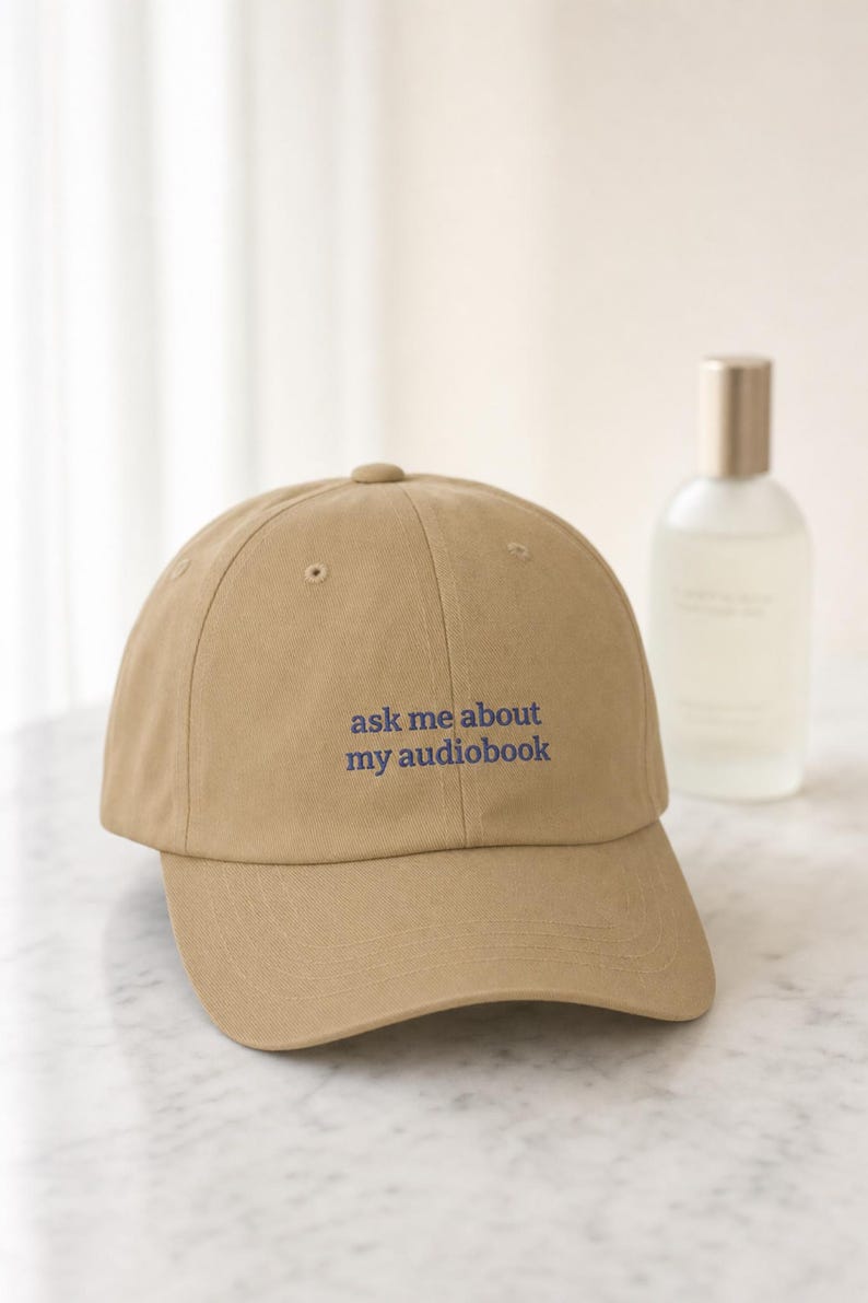 'Ask Me About My Audiobook' embroidered dad hat styled on pale marble beside a frosted glass bottle, shot in bright airy window light with shallow depth of field.