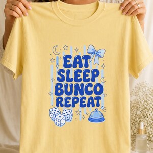 'Eat Sleep Bunco Repeat' bunco dice graphic tee held flat facing camera showing full chest print — bunco gift tee displayed by hands in a warm wellness editorial scene.