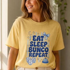 'Eat Sleep Bunco Repeat' bunco dice graphic tee worn by a woman in a warm sunlit interior — bunco lifestyle gift shirt on model, chest print fully legible, editorial wellness portrait.
