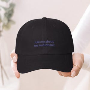 Elegant hands holding 'Ask Me About My Audiobook' embroidered dad hat toward camera, gold ring detail, soft bright ivory background with eucalyptus bokeh.