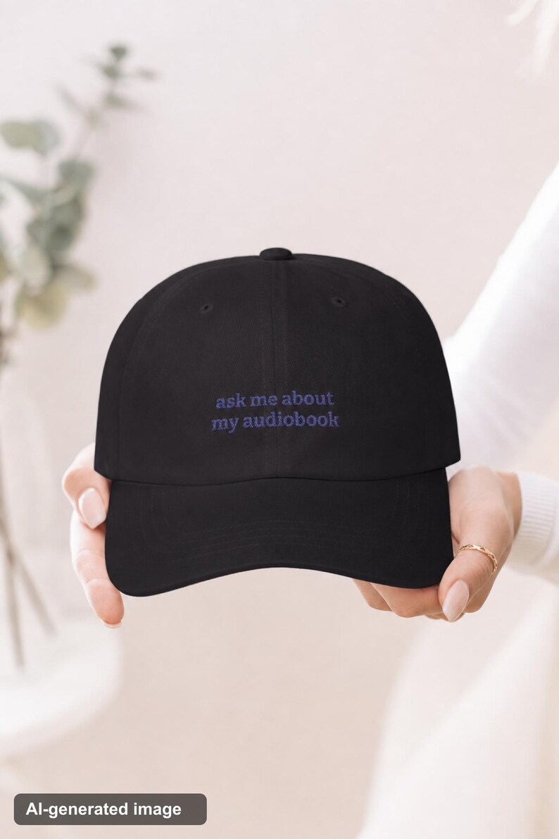 Elegant hands holding 'Ask Me About My Audiobook' embroidered dad hat toward camera, gold ring detail, soft bright ivory background with eucalyptus bokeh.
