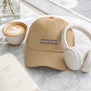 'Ask Me About My Audiobook' dad hat on white marble café table beside a ceramic latte, open book, and white headphones in bright airy natural light — editorial lifestyle moment.