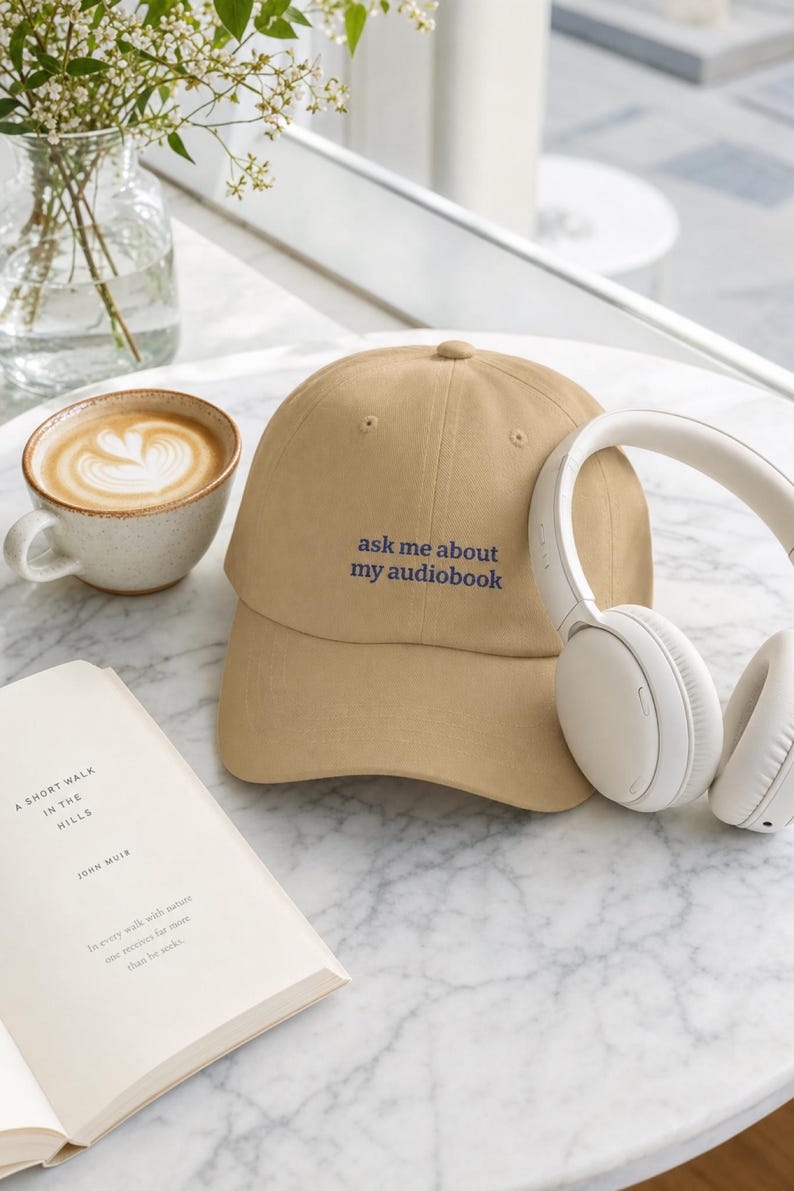 'Ask Me About My Audiobook' dad hat on white marble café table beside a ceramic latte, open book, and white headphones in bright airy natural light — editorial lifestyle moment.
