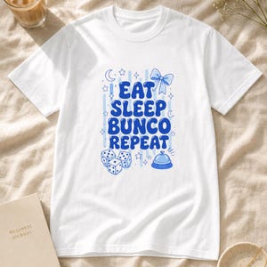 'Eat Sleep Bunco Repeat' printed t-shirt bunco dice graphic tee in a sunlit cream linen flat lay with wellness props — perfect bunco night gift or bunco group tee.
