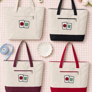 May include: Four canvas tote bags with zippered front pockets, each featuring a graphic of two mahjong tiles. One bag is solid off-white, while the others have contrasting colored bottoms and handles in black, dark purple, and bright red.
