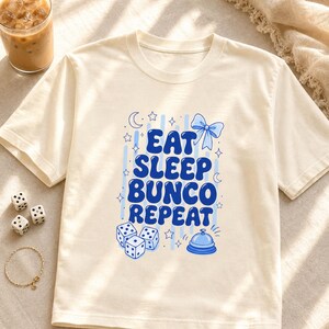 'Eat Sleep Bunco Repeat' bunco dice graphic tee styled in a warm bunco night table scene with dice and wellness props — bunco lifestyle gift shirt, chest print fully visible editorial flat lay.
