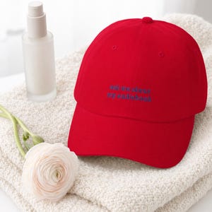'Ask Me About My Audiobook' embroidered dad hat on an ivory bouclé throw with a frosted skincare bottle and soft floral stem, bright luminous window light on white surface.