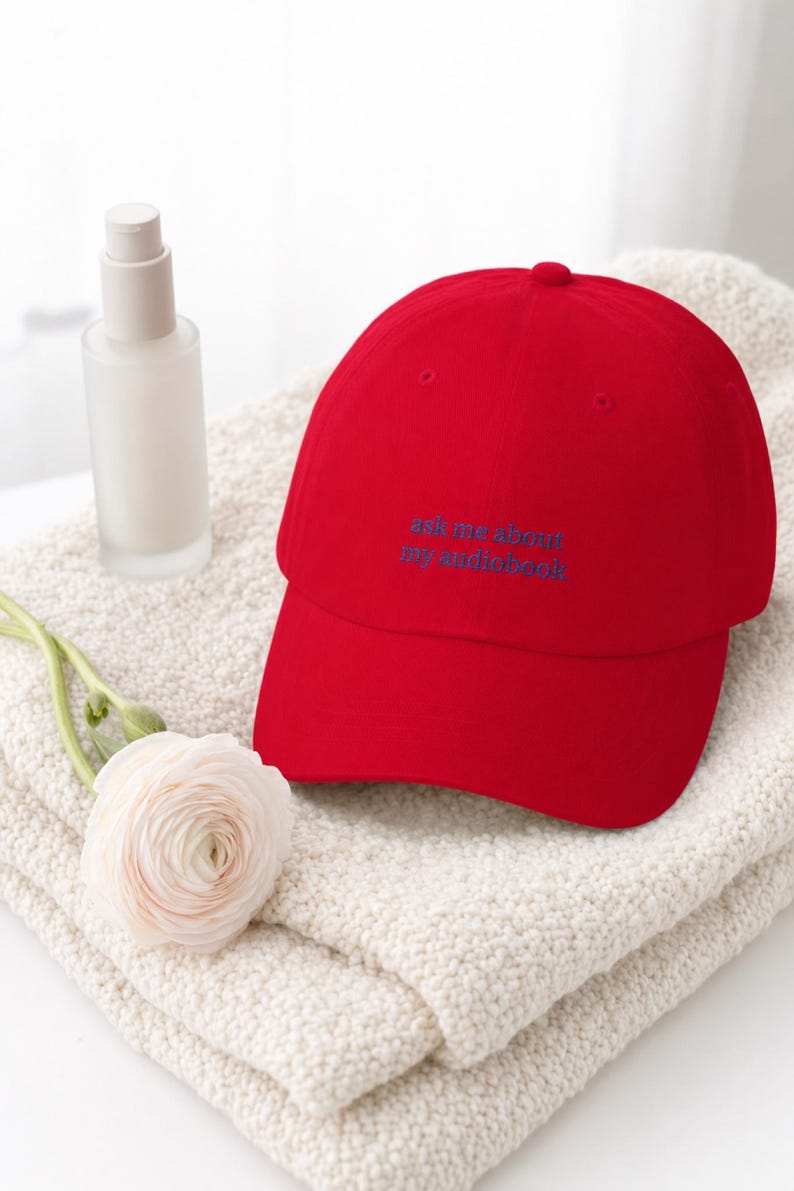 'Ask Me About My Audiobook' embroidered dad hat on an ivory bouclé throw with a frosted skincare bottle and soft floral stem, bright luminous window light on white surface.
