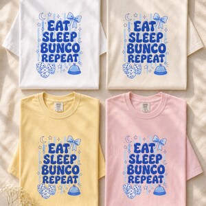 'Eat Sleep Bunco Repeat' bunco dice graphic tee in 4 garment-dyed colorways — bunco lifestyle gift tee color options overhead flat lay, all chest prints legible, editorial wellness scene.