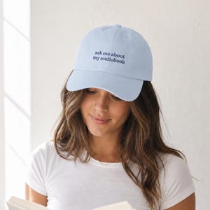 Woman wearing 'Ask Me About My Audiobook' embroidered dad hat in white tee, holding an open book in soft bright window light — candid clean-girl editorial portrait.