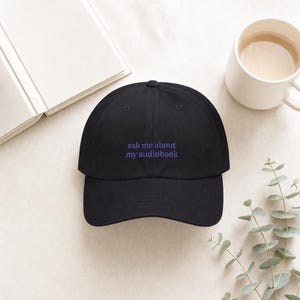 Overhead flat lay of 'Ask Me About My Audiobook' embroidered dad hat on ivory linen beside an open book, white ceramic mug, and eucalyptus sprig in soft natural light.