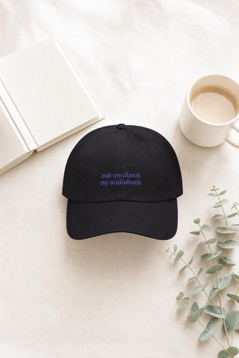 Overhead flat lay of 'Ask Me About My Audiobook' embroidered dad hat on ivory linen beside an open book, white ceramic mug, and eucalyptus sprig in soft natural light.
