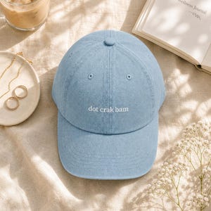 'Dot Crak Bam' embroidered mahjong dad hat — denim adjustable cap, mahjong lover gift. Overhead flat lay on cream linen with iced latte, gold jewelry, and baby's-breath in warm morning light.