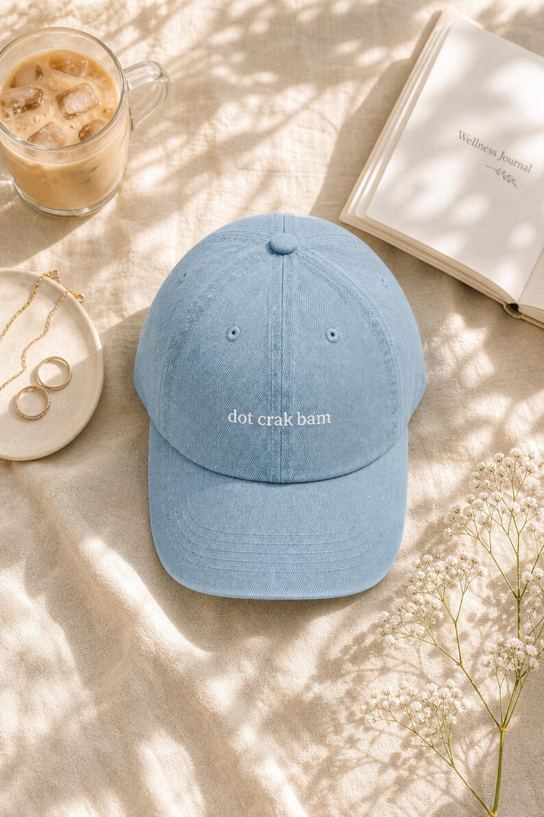 'Dot Crak Bam' embroidered mahjong dad hat — denim adjustable cap, mahjong lover gift. Overhead flat lay on cream linen with iced latte, gold jewelry, and baby's-breath in warm morning light.