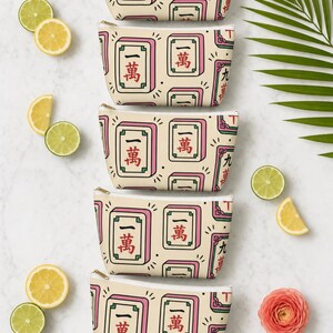 Multiple colorways of the mahjong tile-print accessory pouch in an overhead flat lay on white marble — mahjong gift pouch color options, bright Palm Beach editorial group shot.