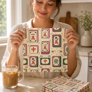 May include: A square fabric napkin featuring a repeating pattern of colourful mahjong tiles on a cream background. The tiles display various Chinese characters and symbols in red, green, and black ink. A stack of identical folded napkins sits nearby.
