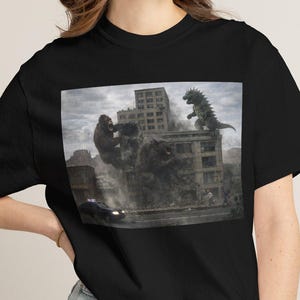 May include: Black t-shirt featuring a graphic of a city scene with Godzilla and King Kong battling. The image shows the monsters destroying buildings, with a car on the road below. The shirt is a classic crew neck style.