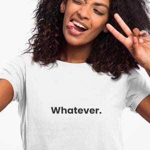 Pode incluir: Uma camiseta branca com a palavra "Whatever." impressa em preto. Uma pessoa está segurando um smartphone preto e fazendo um sinal de paz com os dedos enquanto mostra a língua.