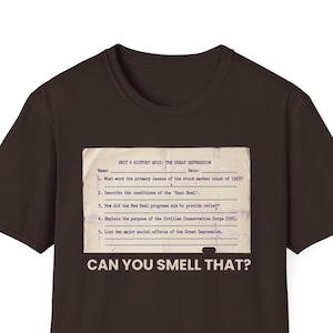 May include: A dark brown t-shirt featuring a vintage-style quiz sheet graphic. The quiz sheet includes questions about the Great Depression and the text "CAN YOU SMELL THAT?" The shirt is a casual wear item.