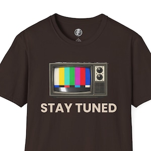 May include: A brown t-shirt featuring a retro television graphic with a colorful test pattern on the screen. The words "STAY TUNED" are printed in white below the television. The shirt has short sleeves and a crew neck.