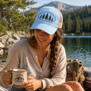 May include: A light blue trucker hat with a black oval design of pine trees. The person is holding a beige mug with the text "Take the Scenic Route" and a mountain graphic. The person is wearing a light beige long-sleeve shirt.