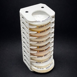 May include: A white, vertical rack holding multiple petri dishes. The dishes are labeled with handwritten text, including "BLUE OYSTER" and "PINK OYSTER". The rack has a slotted design and a curved top with two holes.