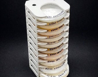 Puede incluir: Un soporte blanco vertical que contiene múltiples placas de Petri. Las placas están etiquetadas con texto escrito a mano, incluyendo "BLUE OYSTER" y "PINK OYSTER". El soporte tiene un diseño ranurado y una parte superior curvada con dos agujeros.