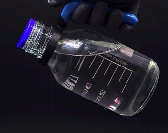 Puede incluir: Una botella de laboratorio transparente y graduada con tapa y asa azules, llena de líquido transparente. La botella tiene marcas negras y la sostiene una mano enguantada sobre un fondo oscuro. La botella mide unos 20 cm de alto.