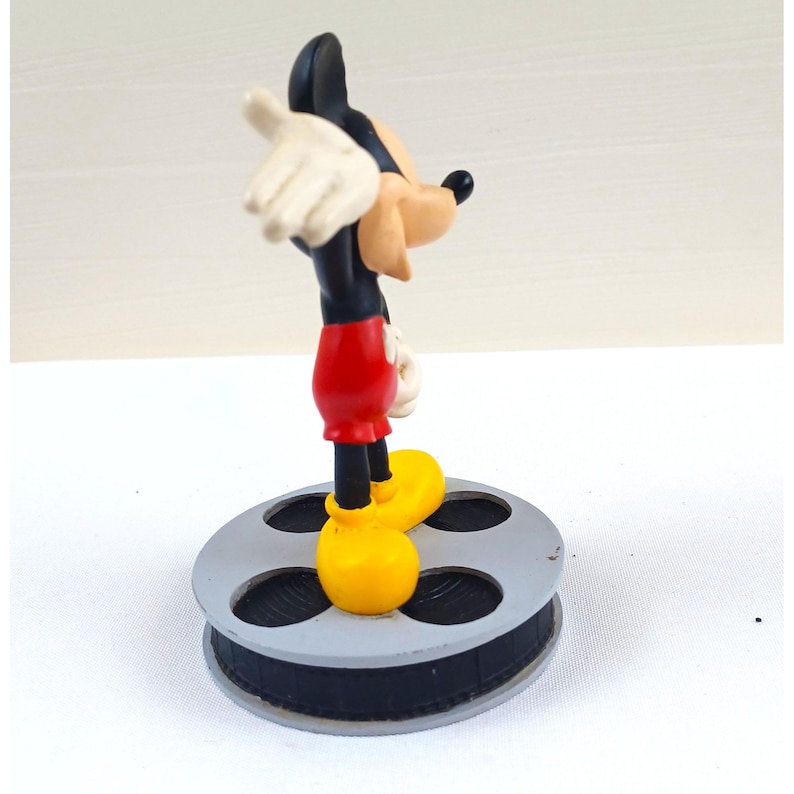 May include: A Mickey Mouse figurine in a classic pose, wearing red shorts, yellow shoes, white gloves, and a black body. The figurine stands on a gray film reel base, a nod to his cinematic history.