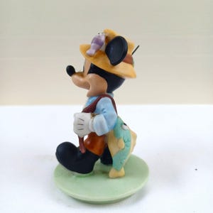 May include: A figurine of Mickey Mouse dressed as a fisherman. He wears a yellow hat, blue shirt, brown overalls, and black shoes. A fish hangs from his back. The figurine stands on a green circular base.