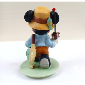 May include: A figurine of Mickey Mouse dressed as a fisherman, viewed from the back. He wears a yellow hat with a brown band, a light blue shirt, orange pants, and black shoes. He holds a fishing rod and a fish. The figurine stands on a green base.